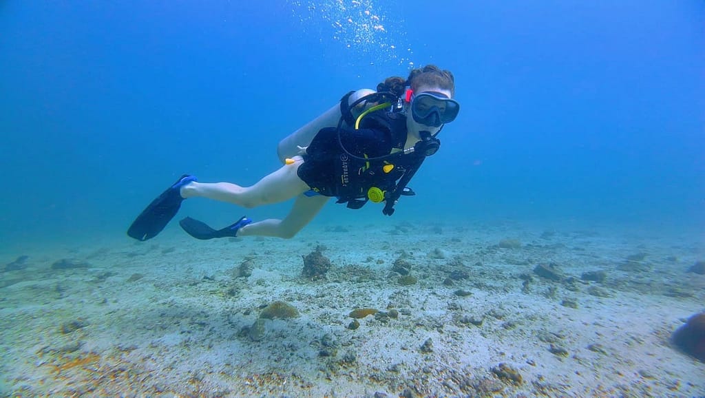 a diver with Buoyancy releasing air bubble, she is really close to the ground underwater, with the sand and blue water 