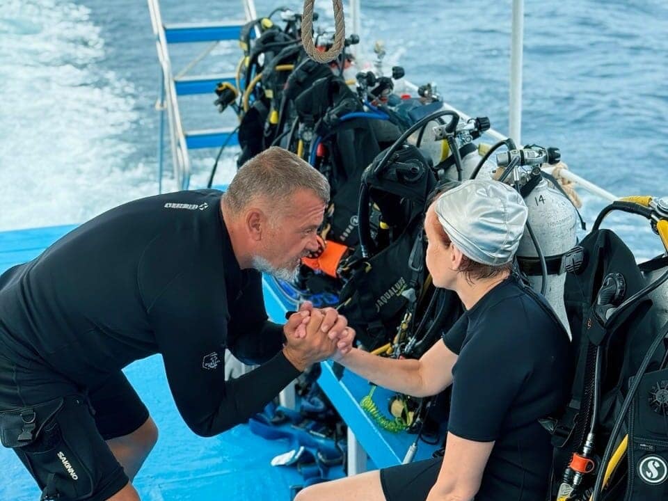 On a boat, with a high amount of scuba diving equipments on the background, a diving instructor is motivating his student for her next step right before diving.